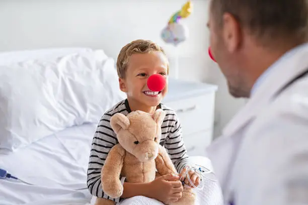 Sick little boy wearing red nose sitting on hospital bed playing with doctor while holding teddy bear. Portrait of cheerful kid with clown nose talking to pediatrician at clinic. Happy hospitalized child in hospital with general practitioner during a clown therapy.