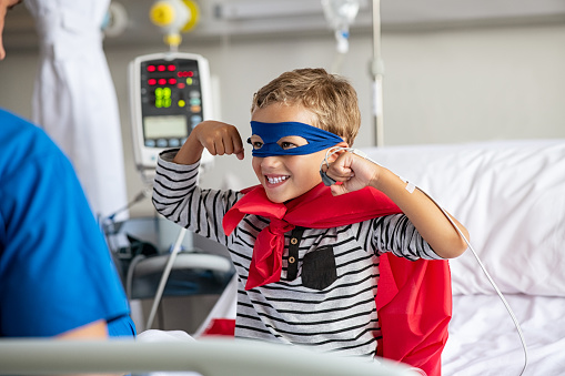 Cheerful strong little boy wearing blue eyeband and red cape like superhero sitting in hospital bed playing with nurse. Playful child gesturing dressed in superhero costume at clinic overcome adversity and health challenge. Happy smiling kid playing at hospital.