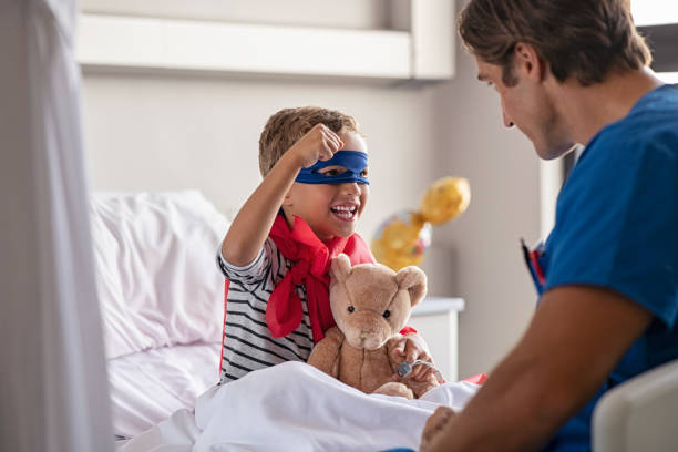 Little cute boy dressed like superhero with nurse Strong little boy wearing superhero blue mask and red cape sitting on hospital bed playing with nurse. Happy superhero kid gesturing with playful doctor at clinic. Happy child playing as superman in pediatric ward with healthcare worker. man with teddy bear stock pictures, royalty-free photos & images