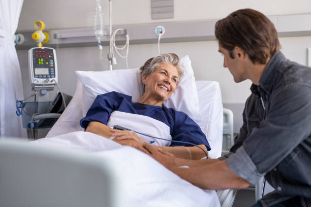 Son visiting his smiling mother at hospital Son visiting sick old mother in hospital. Smiling senior patient in conversation with man. Grandson visiting and cheering his happy granny lying in bed at modern hospital ward. wards stock pictures, royalty-free photos & images