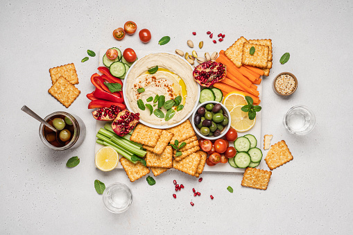 Carrot, cucumber, tomato, celery sticks, dry crackers, olives and hummus bowl on marble tray and light background, top view