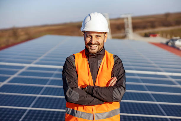 A happy proud worker standing on the roof with solar panels and supporting eco living. A handyman standing on the rooftop with solar panels and smiling at the camera. career vitality stock pictures, royalty-free photos & images
