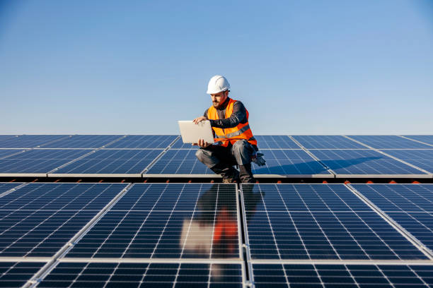 A worker on the roof using laptop charged by sun energy. A handyman looking at the laptop on rooftop and testing solar panels. solar panel stock pictures, royalty-free photos & images