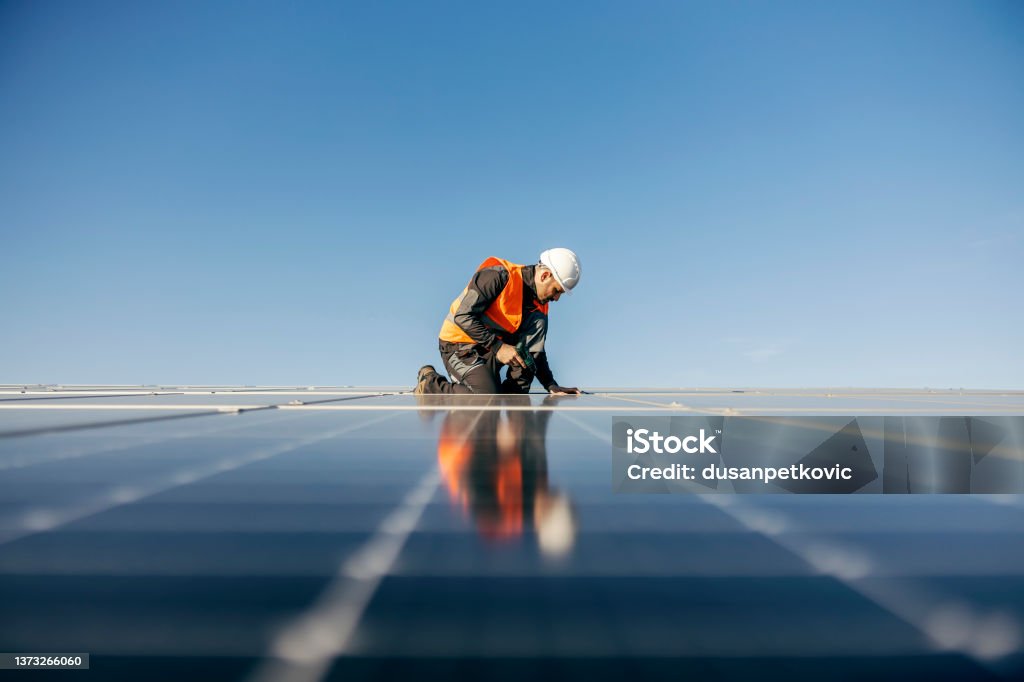 Worker kneeling and sets solar panel. A handyman on the rooftop installing solar panels. Solar Power Station Stock Photo