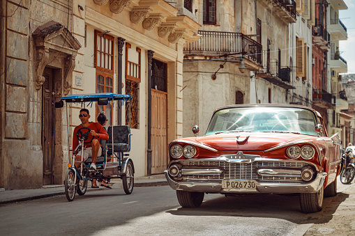 Havana, Cuba - March 22, 2019: Shiny retro car parked on the street and bicycle taxi driver riding by