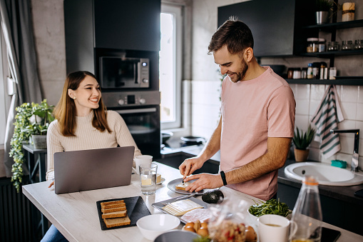 Young woman working from home while her partner is making a breakfast