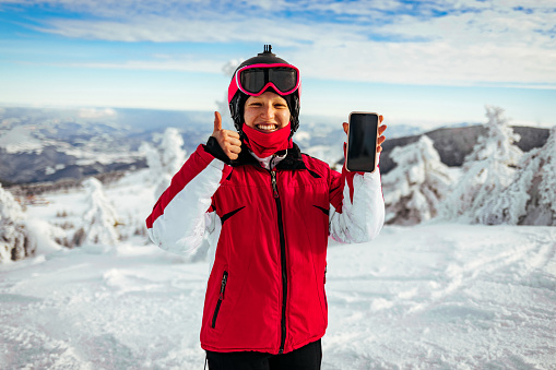 Young skier woman wearing ski goggles holding smartphone showing screen doing ok sign with fingers, excellent symbol.