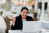 A young Indian woman is holding a video conference sitting in a cafe on the terrace. Freelance, online business.
