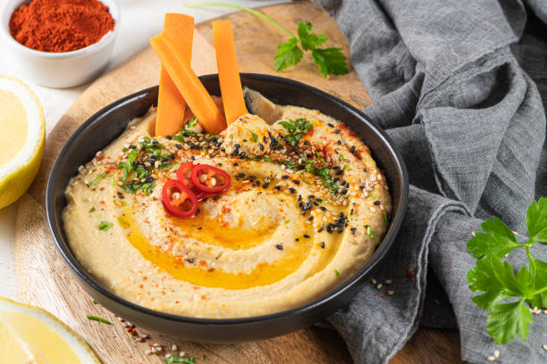 delizioso hummus in fiocco di ceramica con bastoncino di carota immerso nell'hummus. composizione colorata di snack su sfondo bianco. - hummus foto e immagini stock