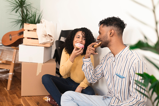 Portrait of a happy, young mixed race couple taking a break and eating donuts while moving into a new home
