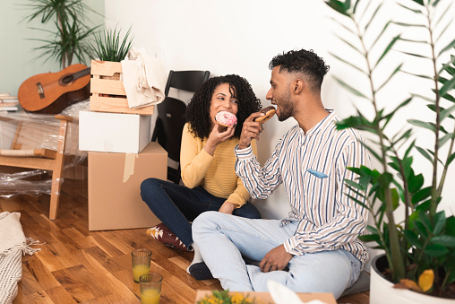 Portrait of a happy, young mixed race couple taking a break and eating donuts while moving into a new home