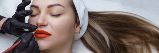 istock banner Young woman with permanent makeup on her lips in a cosmetologists salon. Permanent makeup tattoo. draw a line with a red lip pencil 1373287890