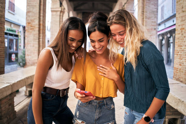 trois femmes heureuses utilisant un téléphone portable à l’extérieur. groupe d’amies souriantes regardant les médias sociaux sur smartphone - jeunes filles photos et images de collection