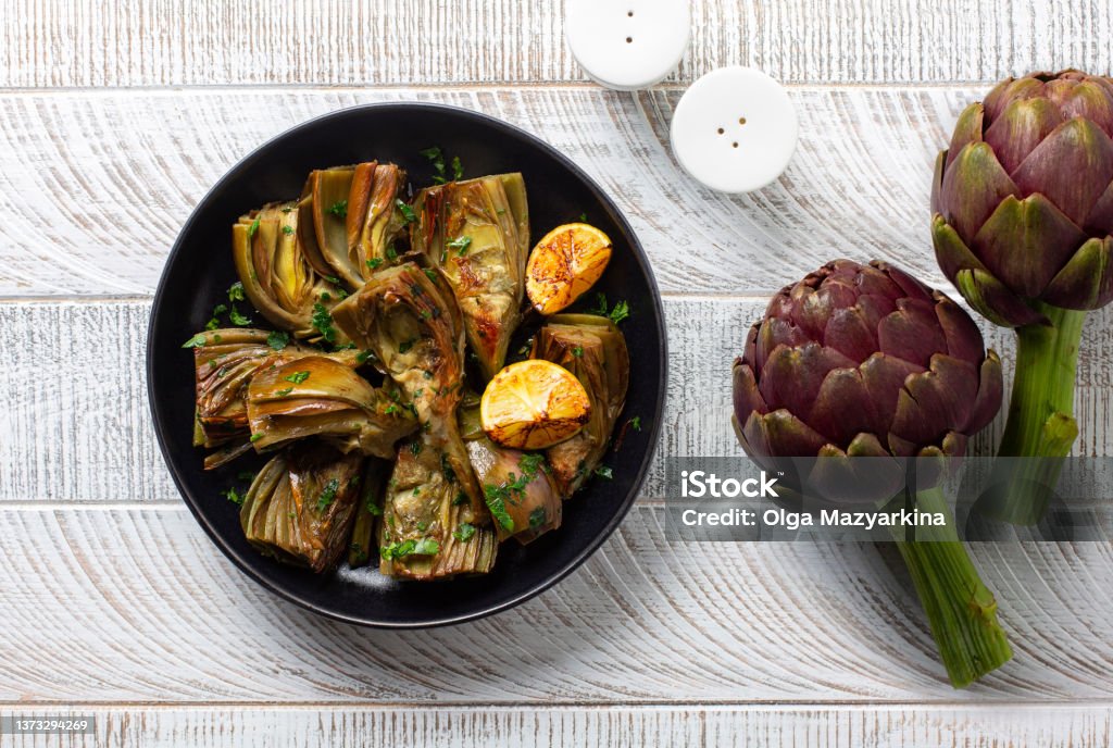 Artichoke or carciofi vegetable, raw and cooked. Artichokes boiled and roasted in olive oil with garlic, garnished with parsley and lemon. White wooden table. Top view. Artichoke Stock Photo