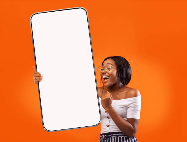 Happy excited teen african american female in glasses rejoices to success and shows big smartphone with blank screen Happy excited teen african american female in glasses rejoices to success and shows big smartphone with blank screen, on orange background, studio. Gestures, great news, application, ad, good site huge black woman pictures stock pictures, royalty-free photos & images