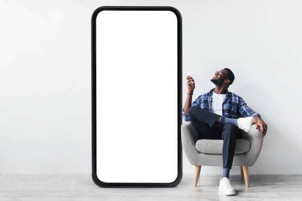 istock Smart home system. Young black guy with remote control sitting in armchair near huge smartphone, mockup for design 1373299907
