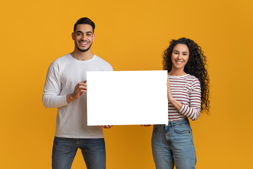 Ad Mockup. Happy Young Arab Spouses Holding White Blank Placard In Hands, Smiling Middle Eastern Couple Demonstrating Copy Space For Your Design While Standing Over Yellow Studio Background