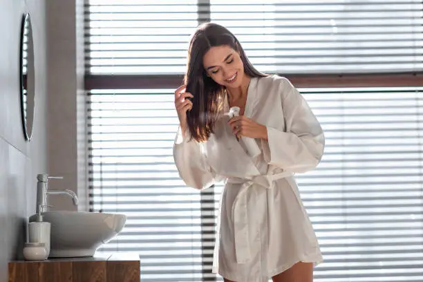 Haircare Cosmetic. Beautiful Millennial Female Applying Moisturising Hair Spray On Dry Ends, Happy Young Woman Wearing White Silk Robe Standing Near Mirror In Bathroom, Making Beauty Routine At Home