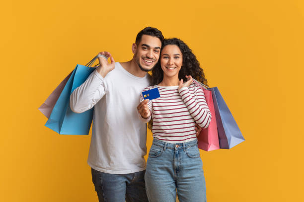 Easy Shopping. Happy Middle Eastern Couple Holding Credit Card And Shopper Bags Easy Shopping Concept. Happy Middle Eastern Couple Holding Credit Card And Bright Shopper Bags, Cheerful Young Arabic Spouses Enjoying Seasonal Sales, Standing Over Yellow Background, Copy Space credit card purchase stock pictures, royalty-free photos & images