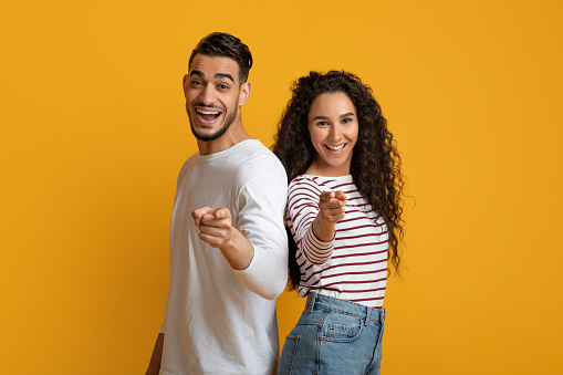 Joyful young arabic couple pointing at camera with fingers, cheerful middle eastern man and woman having fun while indicating somebody, posing together over yellow studio background, copy space