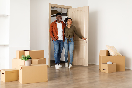 Real Estate Purchase. Happy African American Couple Entering Their New Home Standing Among Cardboard Moving Boxes In Empty Living Room Indoor. Housing For Young Family, Apartment Rent And Ownership