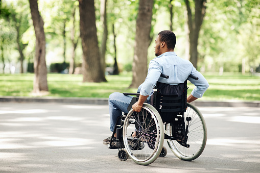 Thoughtful young black guy in wheelchair having walk at urban park, copy space. Millennial African American man with physical disability spending time outdoors, enjoying nature