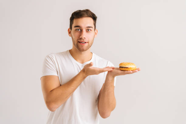 잘 생긴 젊은 남자의 초상화는 카메라를보고, 흰색 고립 된 배경에 맛있는 햄버거를 가리키는. - hamburger bun bread isolated 뉴스 사진 이미지