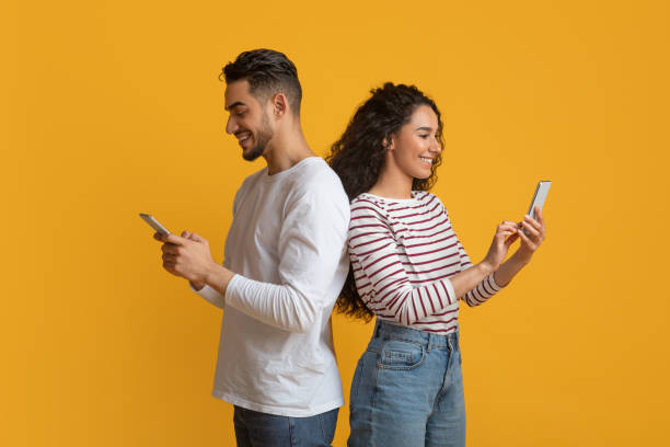 Gadget Addiction. Young Arab Couple Holding Smartphones And Standing Back To Back Gadget Addiction. Young Arab Couple Holding Smartphones And Standing Back To Back, Happy Middle Eastern Man And Woman Busy With Cellphones While Standing Together On Yellow Background, Copy Space excited woman yellow stock pictures, royalty-free photos & images