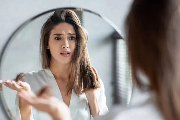 Portrait of stressed young woman with bunch of fallen hair in hand looking at mirror in bathroom, scared upset millennial lady in white silk robe suffering hairloss problem, selective focus