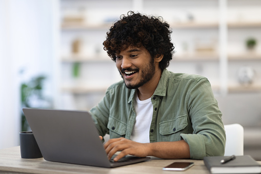 Remote job concept. Happy dark-skinned curly bearded young man in casual sitting at workdesk at home, using modern laptop, typing on computer keyboard and smiling, looking for job online, copy space