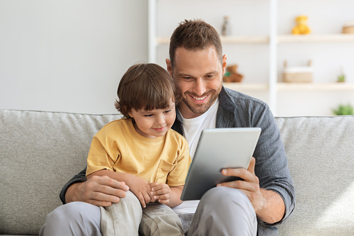 Child development content. Young positive father showing digital tablet to his cute little son, kid enjoying educational show online, sitting on sofa ta home