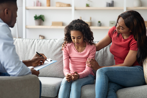 Curly black girl teenager crying during therapy session with male child psychologist, african american mother and daughter visiting psychotherapist together, mom comforting upset kid, clinic interior