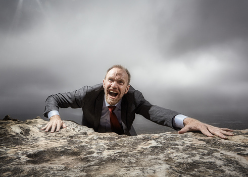Man in a suit looks stressed as he reaches the top of a rocky mountain.