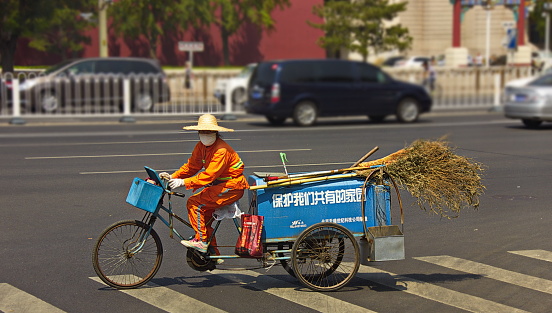 Beijing, China - August 12,2015 : A street cleaner in orange uniform and face mask is crossing the road on tricycle, carrying a traditional bamboo broom.