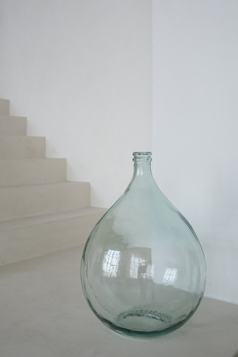 Big drop shaped glass vase standing on the floor of contemporary stylish living room of white color against staircase and wall