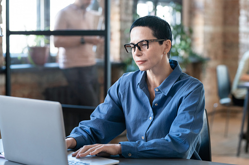 Serious businesswoman with short hair in eyeglasses typing on laptop while sitting at his workplace at office