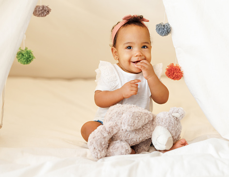 Portrait of cute adorable african american little baby girl infant wearing pink bow headband sitting in teepee play tent at home. Baby development, children and childcare concept