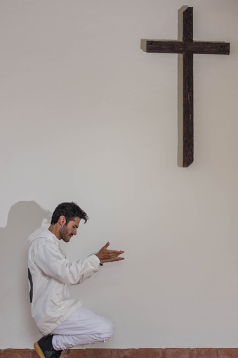 Man dressed in white praying next to a wooden cross in white background