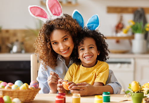 Feliz familia afroamericana: madre enseñando a niño feliz pronto a decorar huevos de Pascua mientras está sentado en la cocina photo