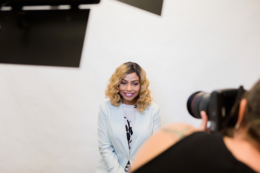 The mid adult female television host smiles as the unrecognizable female photographer takes publicity photos for the studio.