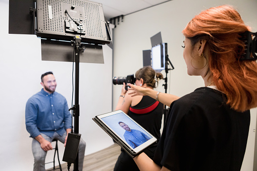 In a studio, the unrecognizable female photographer shoots the television host's headshots as the young adult assistant holding a digital tablet gives directions.  The man's photos are displayed on the tablet screen.