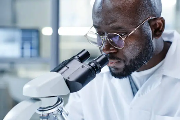 Close-up of African man in eyeglasses looking through the microscope during scientific research in the lab