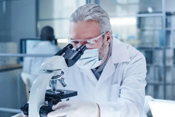 Mature scientist in mask looking through the microscope during scientific research in the laboratory, he working with samples of virus