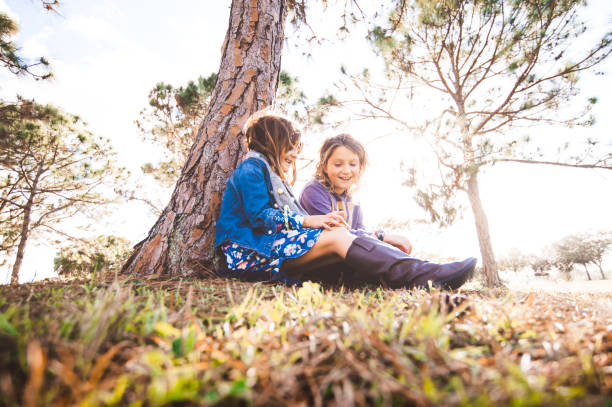 istock Two girls, female friendship, sitting under a tree outdoors laughing with sun flare using a cell phone, technology 1373512258