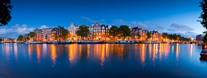 Night time illuminations of pretty doll house apartments and house boats in Amsterdam. Blauwbrug bridge and Munttorren church visible either side. Stitched panoramic image.