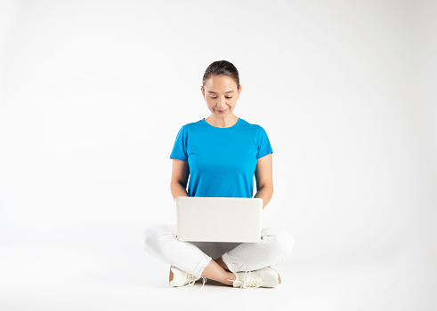Happy beautiful woman sitting on the floor with crossed legs while using laptop on white background.