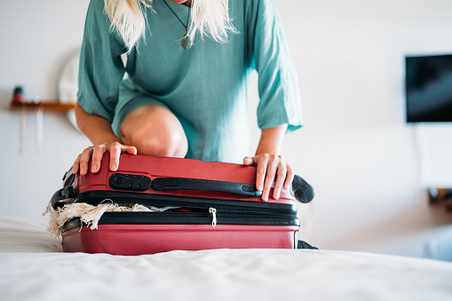 Close-up of a woman in a turquoise dress, trying to close a full suitcase with her knee.