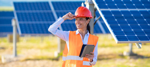 une ingénieure ou une technicienne en électricité portant un gilet orange et un casque de sécurité se tient debout tenant un bloc-notes et souriant tout en travaillant à l’extérieur avec le panneau solaire - solar panel engineer solar power station women photos et images de collection