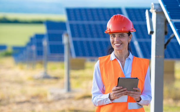 istock A female engineer or electrical technician wearing an orange vest and hard hat stands holding a notepad and smiling while working outdoors with the solar panel 1373564276