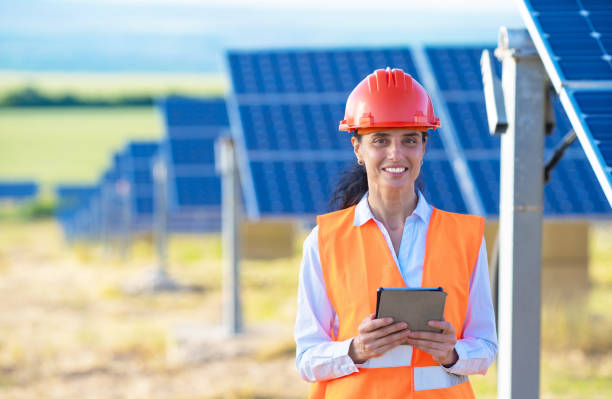 istock A female engineer or electrical technician wearing an orange vest and hard hat stands holding a notepad and smiling while working outdoors with the solar panel 1373564280